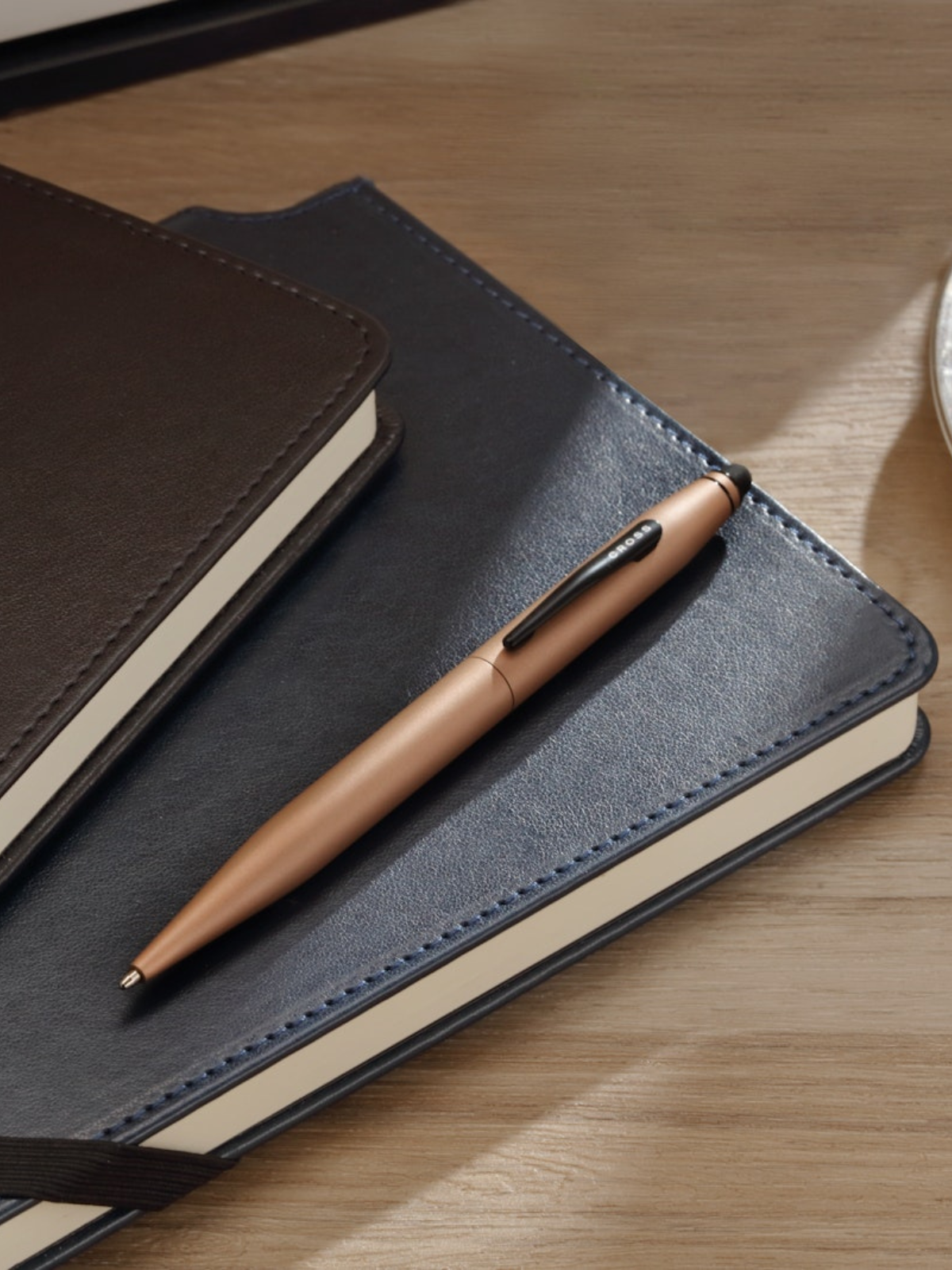 Rose gold pen on a black leather notebook with another notebook in the background on a wooden surface.